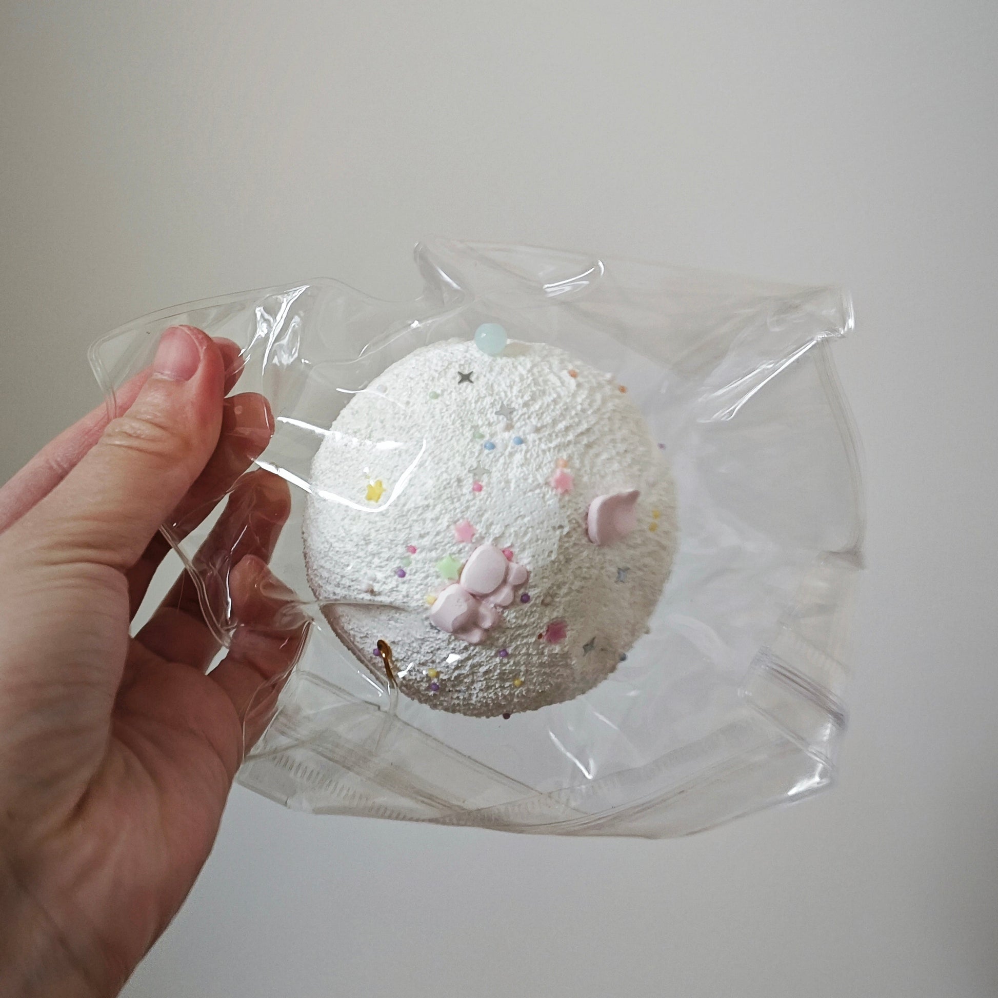 A hand holding a clear plastic bag containing a round, textured white item with small pastel speckles and pink paw-print details, against a plain light gray background.