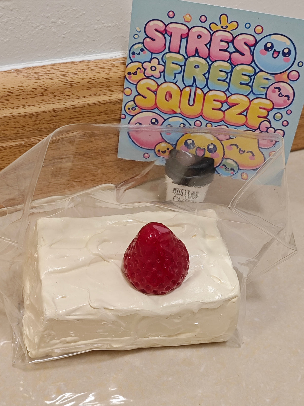 A squishy foam toy shaped like a strawberry-topped cream cake, sealed in a clear plastic bag with a small bottle of glitter and a colorful 'STRESS FREE SQUEEZE' card in the background.