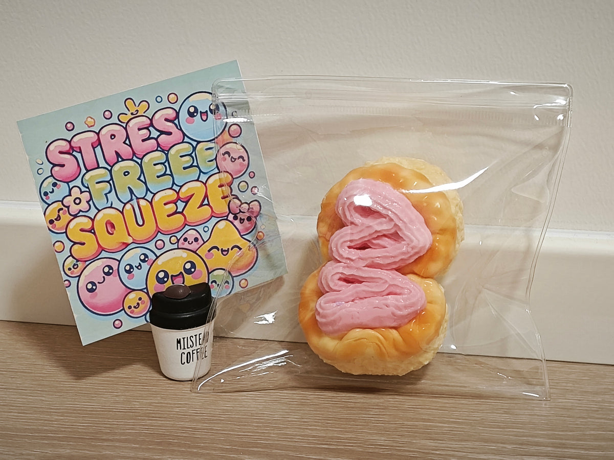 A novelty squishy toy shaped like a pastry with pink frosting, sealed in a clear plastic bag next to a small coffee cup and a colorful 'Stress Free Squeeze' card on a wooden surface.