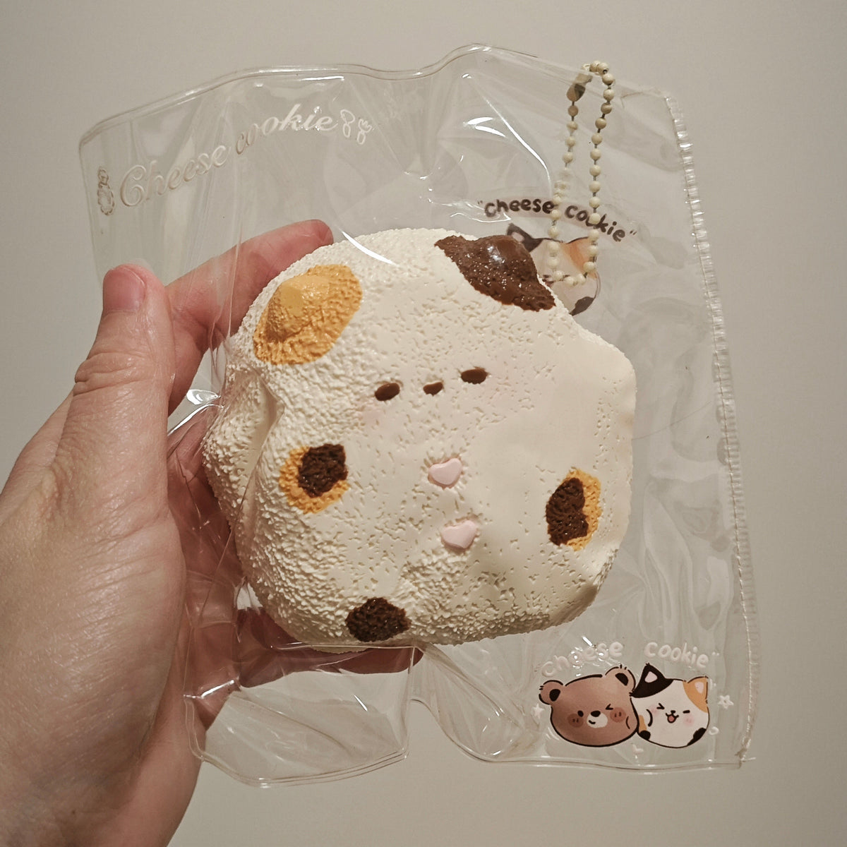 A hand holding a round, soft plush toy shaped like a cow face, still inside a clear plastic bag with printed branding and a small chain attached.