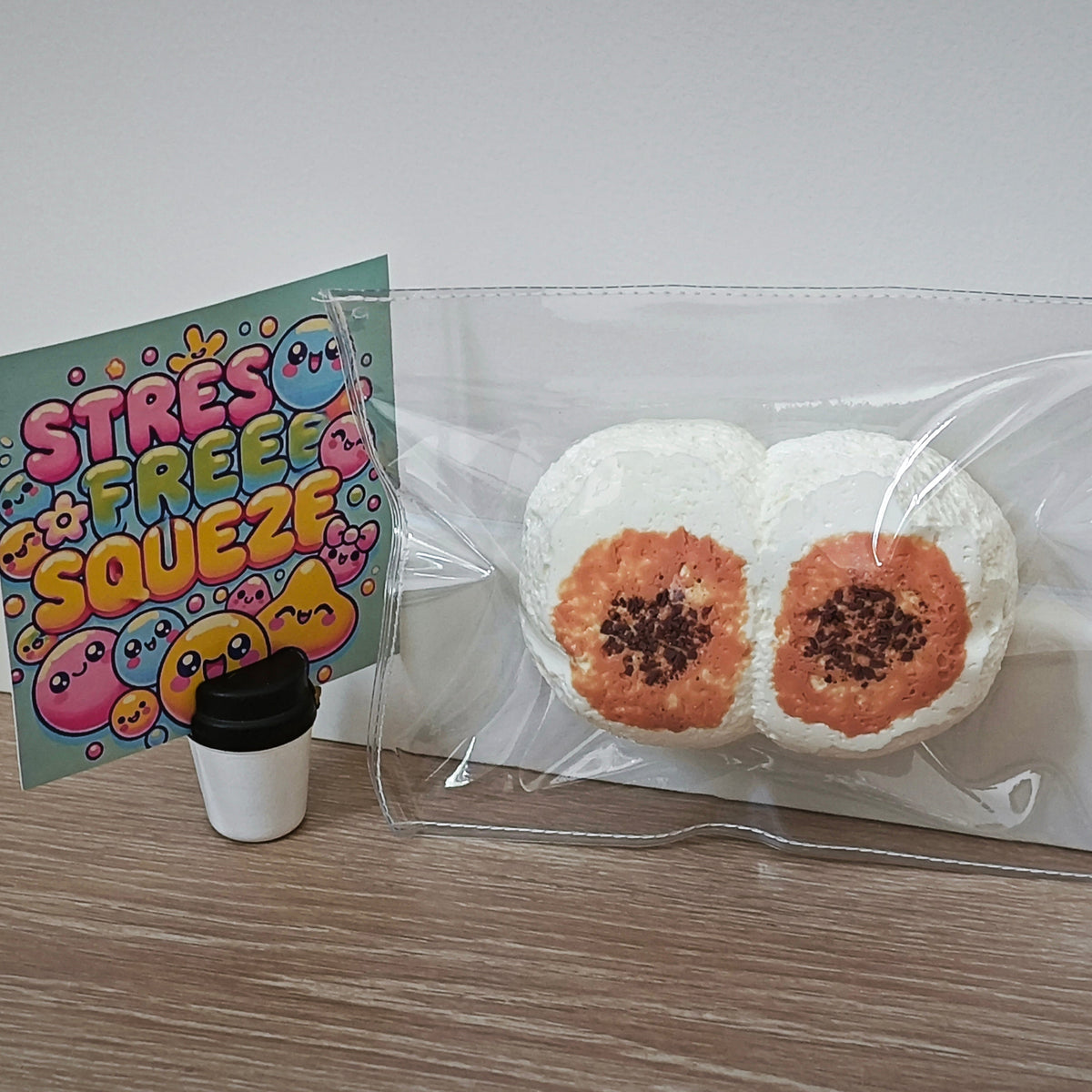 A clear plastic bag containing two white round items with orange centers and dark sprinkles, placed on a wood surface against a white wall. A colorful card reading 'STRESS FREE SQUEEZ' and a small black-and-white cup are also visible beside the bag.
