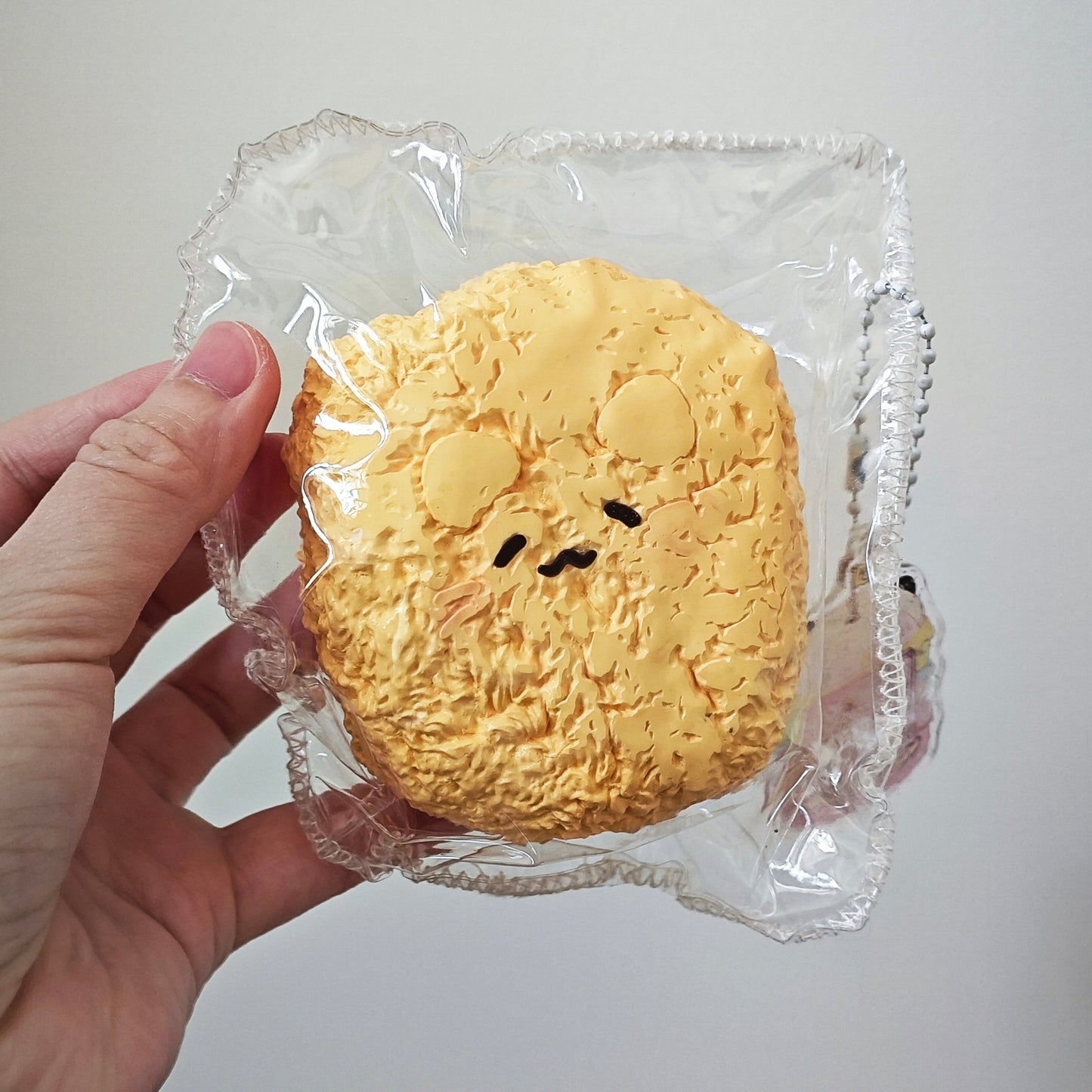 A hand holding a packaged yellow textured toy or sponge shaped like a round character face, sealed in clear plastic against a plain light background.