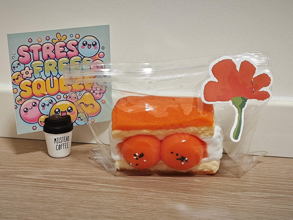 A cute, multi-piece stress toy set featuring a sandwich slice with orange jam and red jam centers, a small cup labeled 'Musten Coffee', and a card that reads 'STRESS FREE SQUEEZE' with cartoon characters. The items are packaged in a clear ziplock bag with a flower sticker on the side, set on a wooden tabletop against a plain wall.