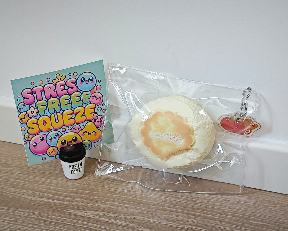 A round beige and orange cookie-themed squishy toy in a clear plastic bag, shown next to a colorful 'STRESS FREE SQUEEZE' card and a small black-and-white 'MISTYAR COFFEE' cup on a wooden surface against a white wall.