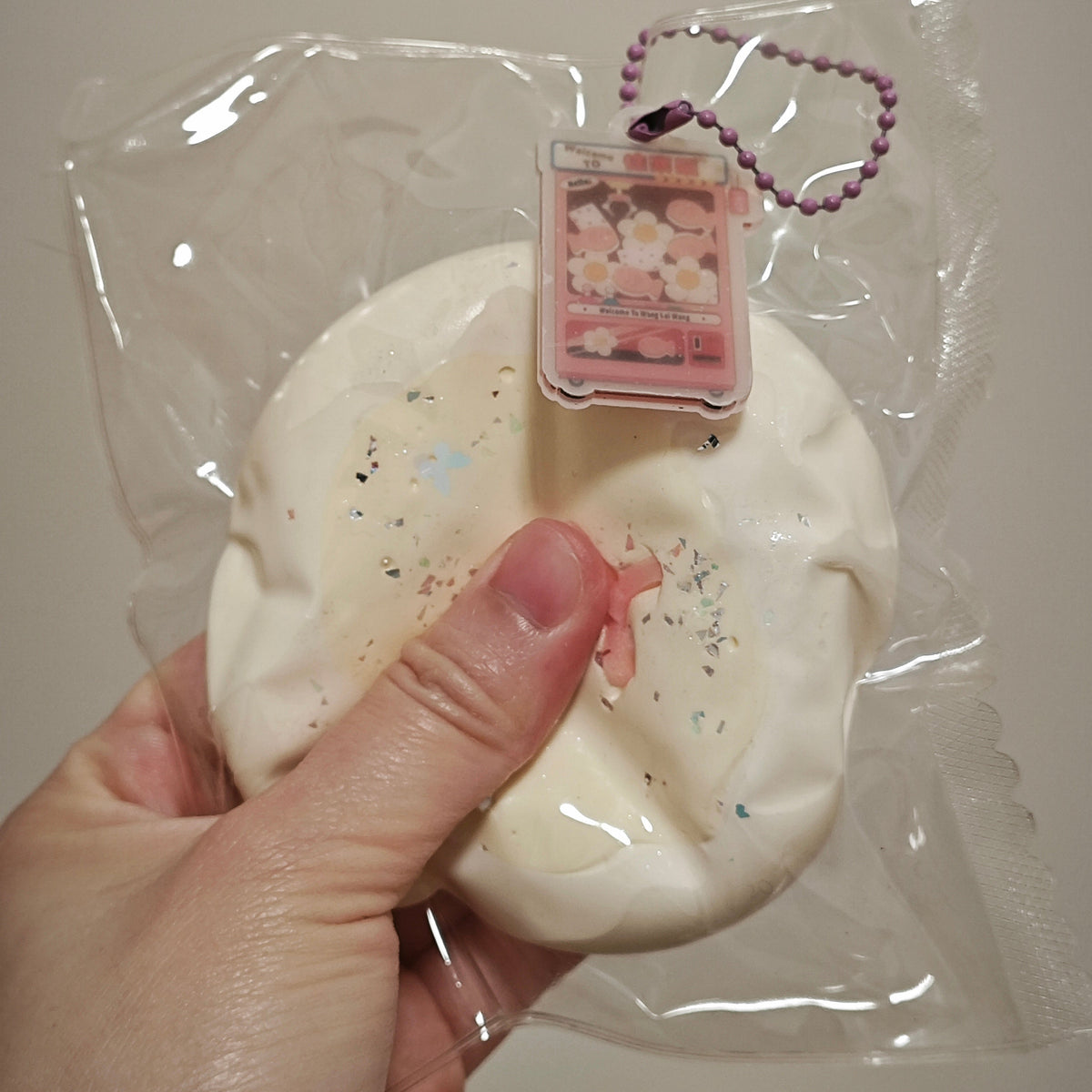 A hand holding a packaged white squishy toy with a small pink charm attached, wrapped in clear plastic against a plain light background.