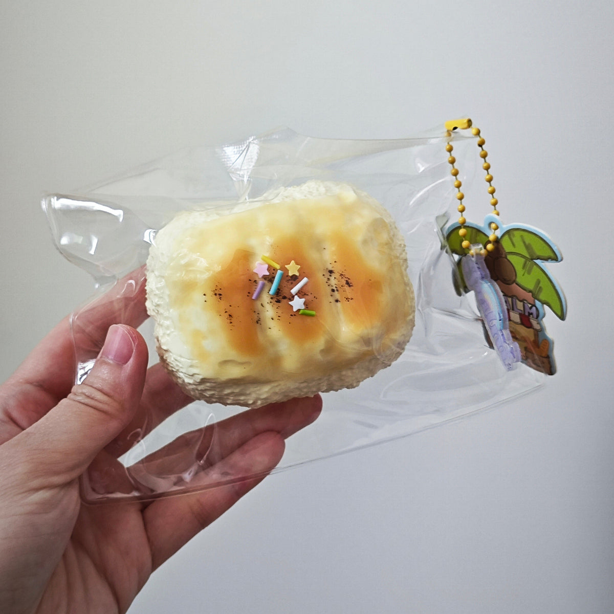 A hand holds a packaged novelty food item shaped like a toasted sandwich or pastry with a small hanging keychain charm, shown against a plain light gray background.