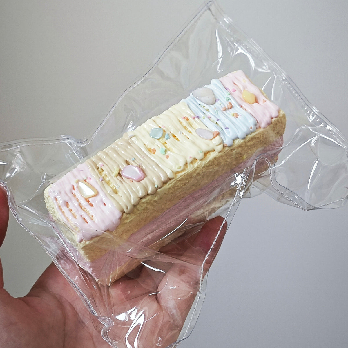 A hand holding a small rectangular cake or pastry wrapped in clear plastic, with pastel pink, cream, and light blue frosting and small decorative candies on top, against a plain light gray background.