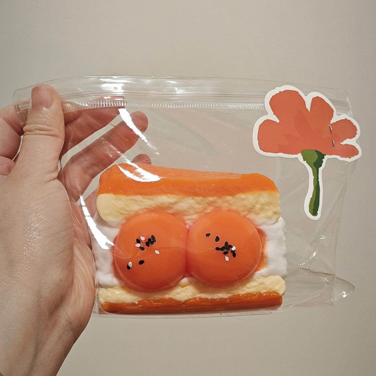 A hand holds a clear plastic zip pouch containing a small orange and cream sandwich-shaped item with two round orange centers, alongside a flower sticker on a plain beige background.