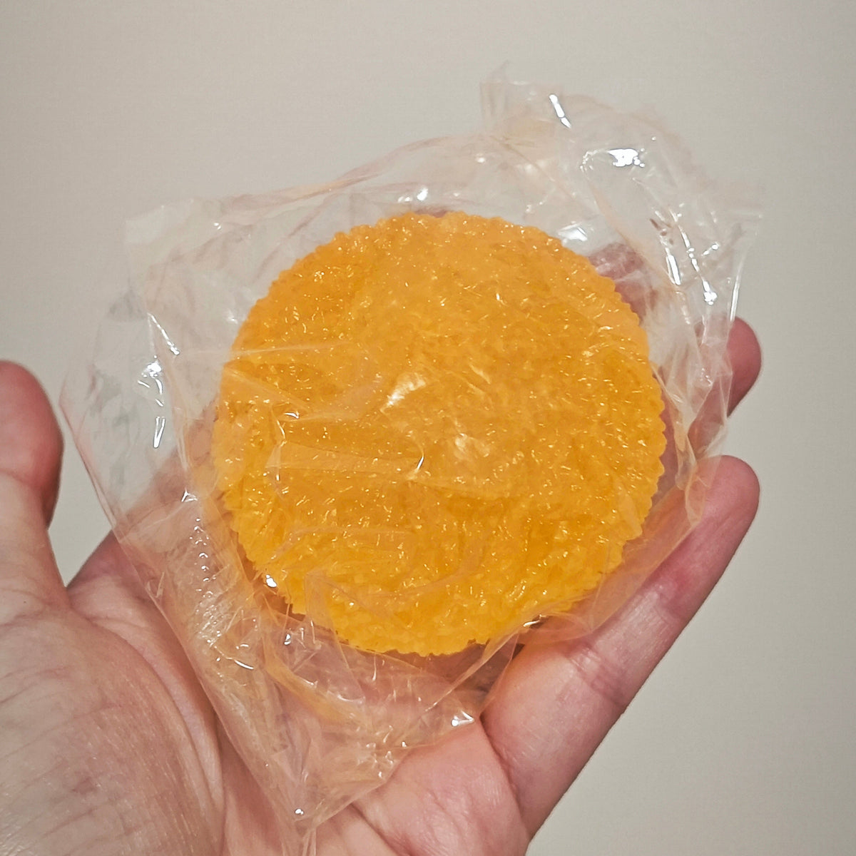 A hand holding a round, textured orange-yellow sponge wrapped in clear plastic against a plain light beige background.