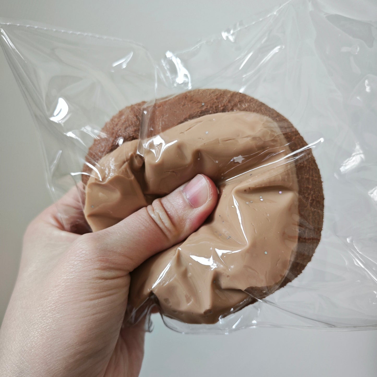 A hand holding a round brown item wrapped in clear plastic, with a tan crumpled paper or fabric insert in the center, against a plain indoor wall.