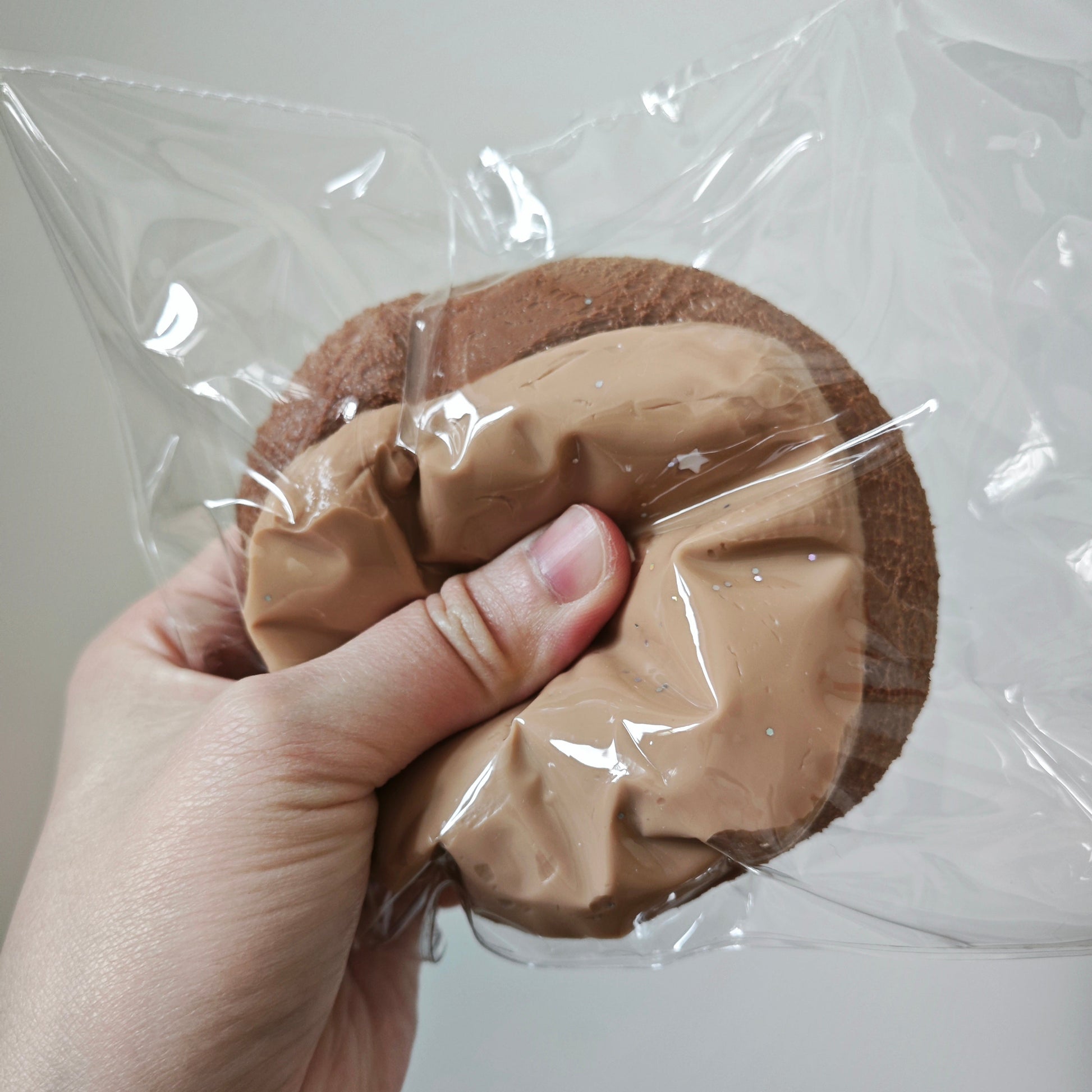A hand holding a round brown item wrapped in clear plastic, with a tan crumpled paper or fabric insert in the center, against a plain indoor wall.