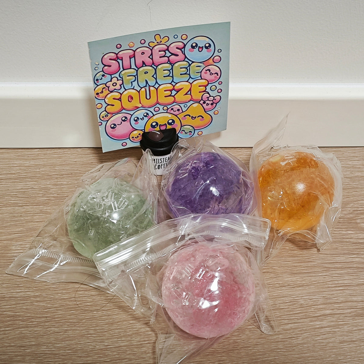 Four colorful stress balls in clear plastic bags arranged on a wooden surface, with a card that reads 'STRESS FREE SQUEEZE' standing behind them.
