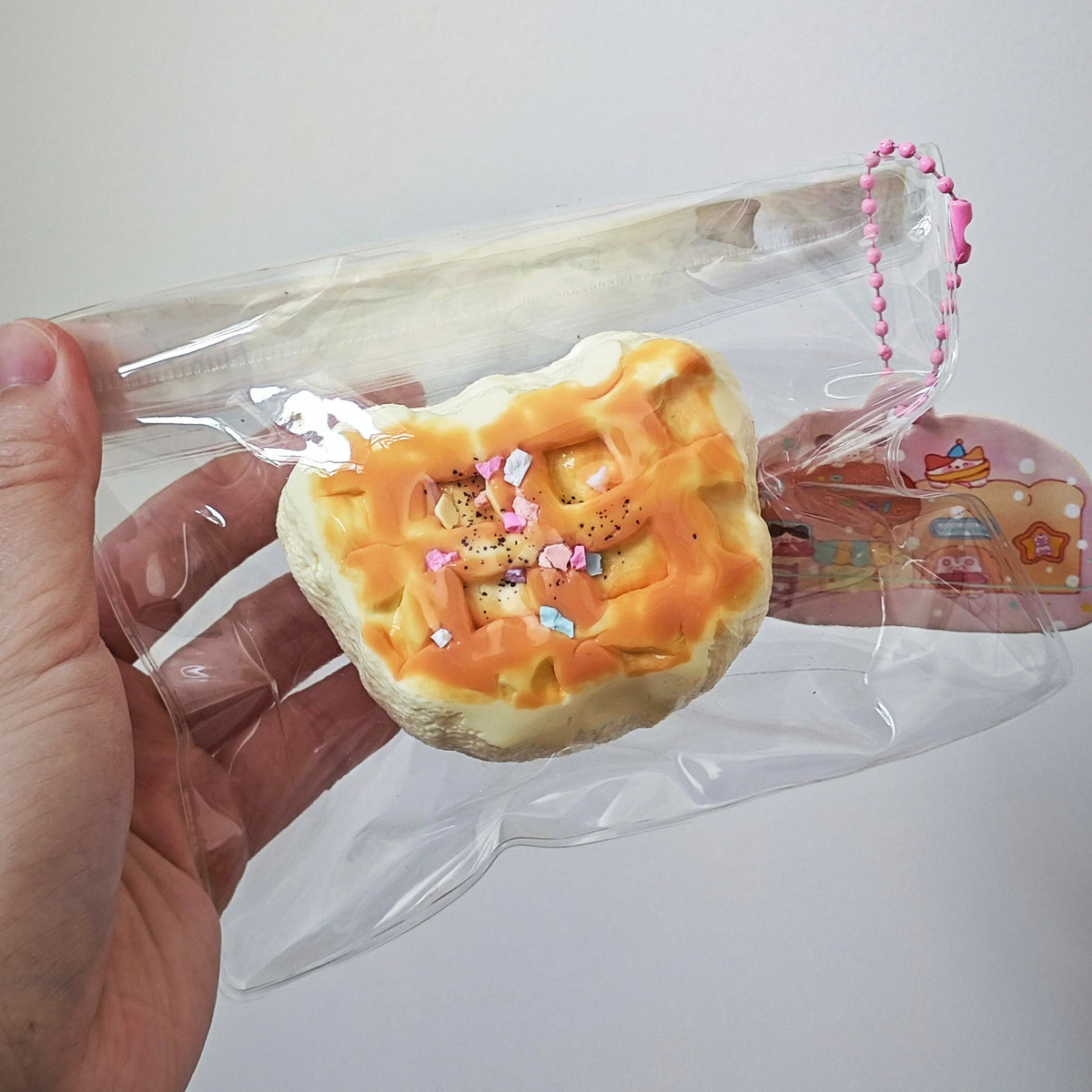 A hand holding a clear plastic bag containing a small, round, waffle-like pastry or bread decoration with orange frosting and pink sprinkles, against a plain light background.