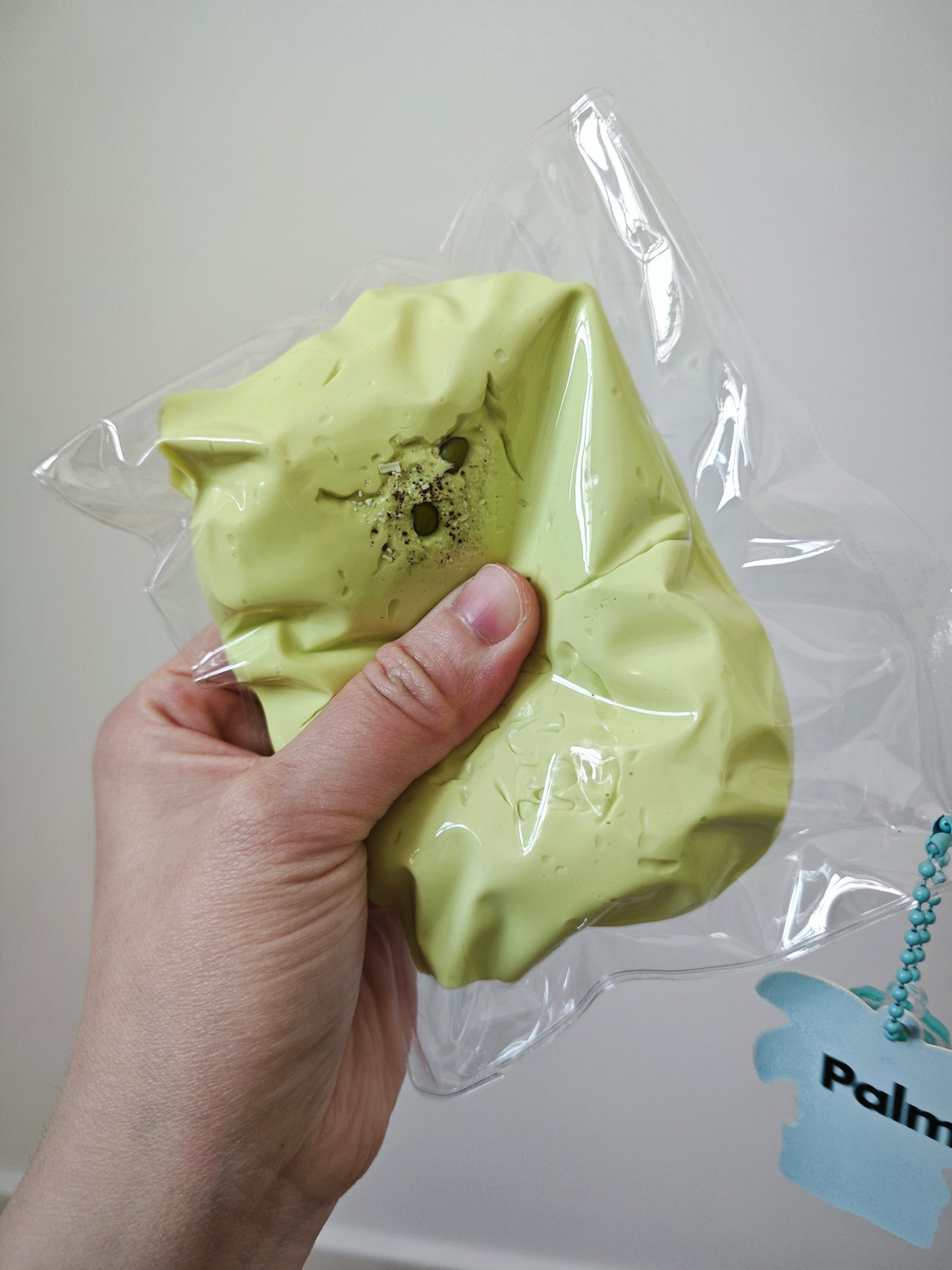 A hand holding a lime green squishy toy inside a clear plastic bag with a light blue tag that reads 'Palm'.
