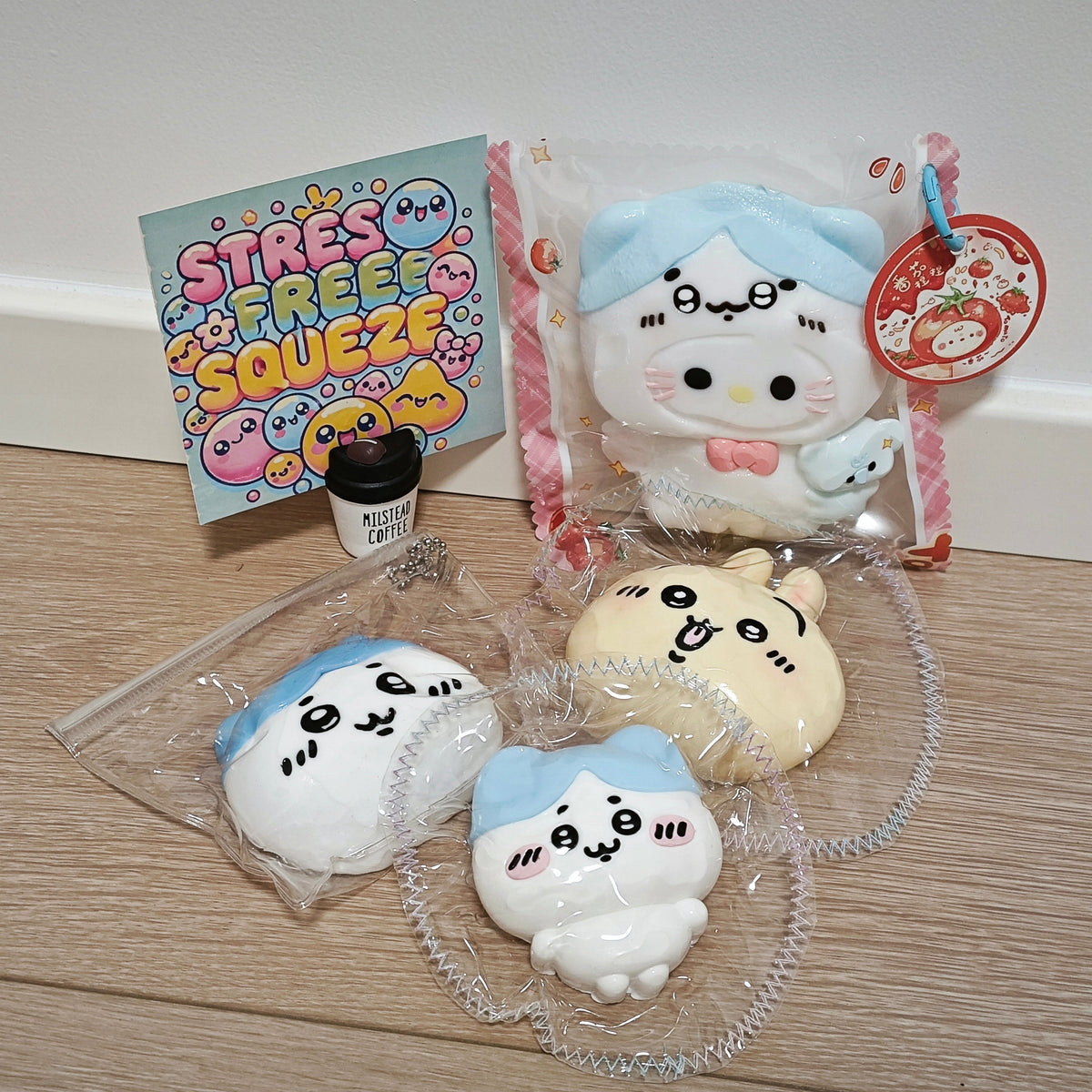 A set of cute cartoon squishies and accessories on a light wood floor against a white wall. The set includes a large plush toy in a clear bag, three smaller squishies in clear bags, a small black cup, and a colorful card that reads 'STRESS FREE SQUEEZE'.