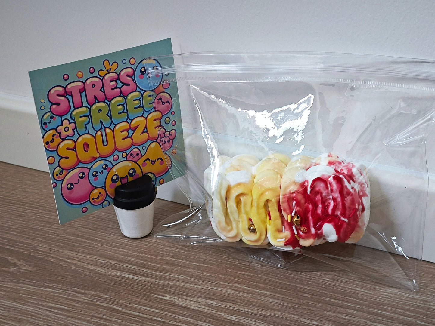 A clear plastic bag containing three novelty squishy toys shaped like a coffee cup, a yellow omelet, and a red-and-white dessert, placed on a wood surface next to a colorful 'STRESS FREE SQUEEZE' card.