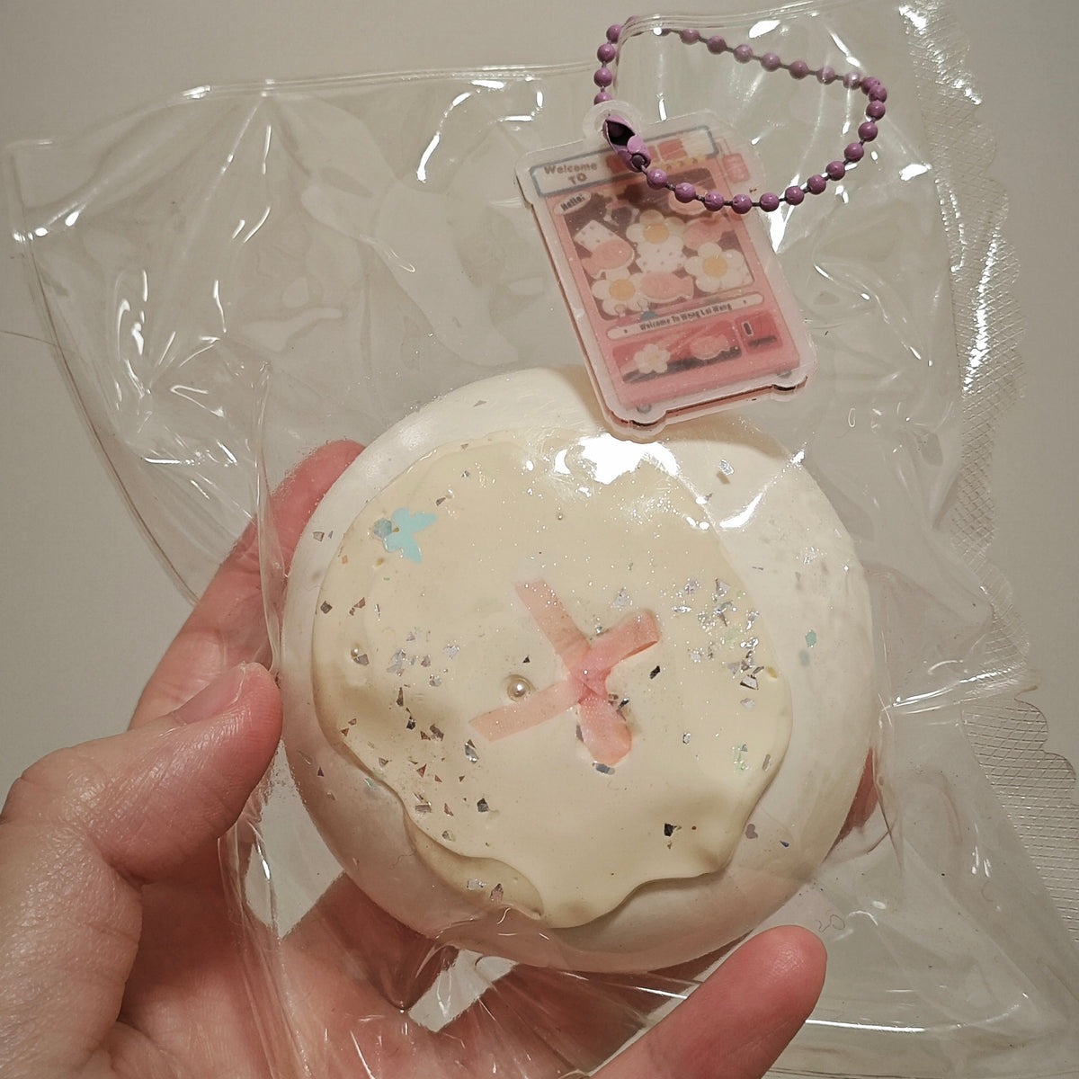 A hand holding a packaged round squishy toy. The toy is off-white with a pink cross design, small pastel blue and black speckles, and a pink bead keychain attached.