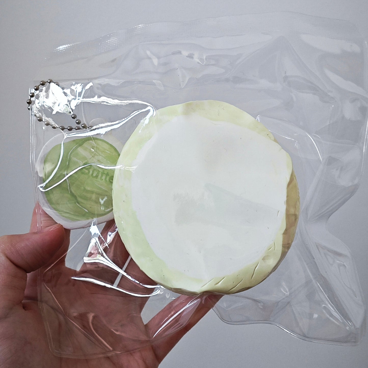 A hand holding a clear plastic bag containing a round, pale yellow and white squishy toy, with a smaller green round accessory attached by a silver ball chain, against a plain light gray background.
