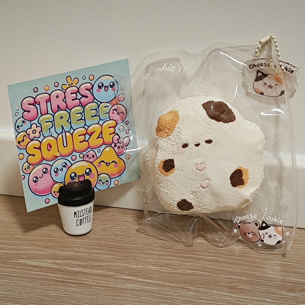 A cute white and brown cow-themed squishy toy in clear packaging, shown with a small 'Mister Coffee' cup accessory and a colorful 'Stress Free Squeeze' card on a wooden surface against a white wall.