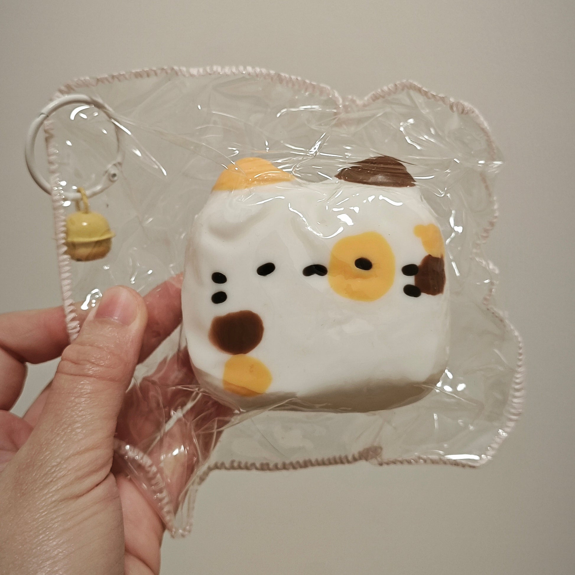 A hand holding a small squishy toy in clear plastic packaging against a plain light beige background. The toy is white with yellow, brown, and black facial markings and a small yellow keychain charm attached.
