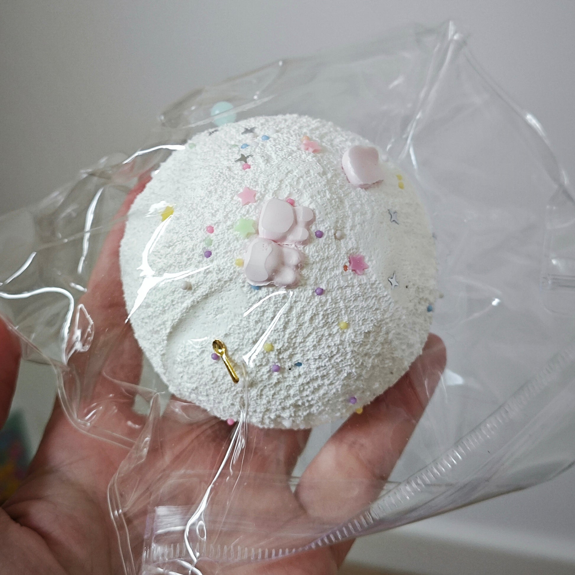 A hand holding a round, textured white item wrapped in clear plastic, decorated with small pastel pink and multicolor confetti-like specks and a small pink bunny detail.