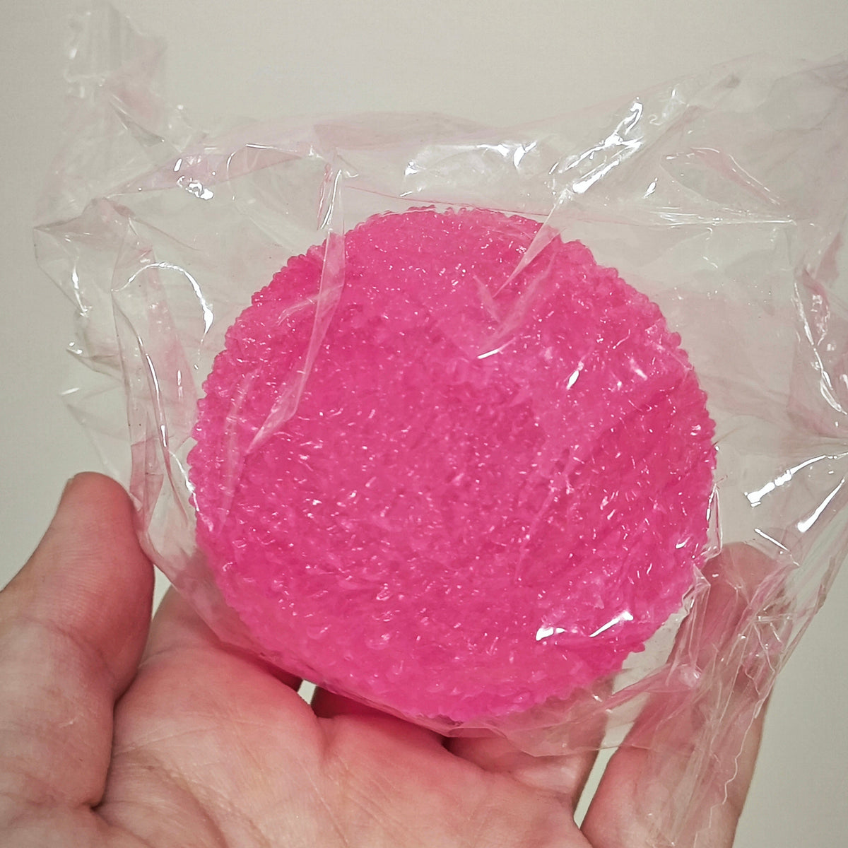A bright pink textured circular sponge or scrubber wrapped in clear plastic, held in a person's hand against a plain light background.