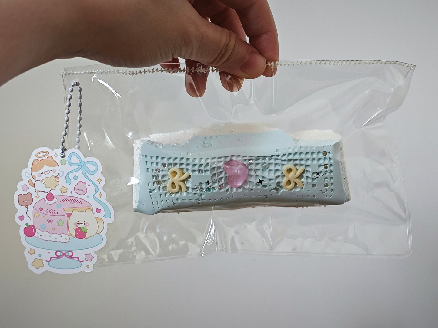 A hand holds a clear plastic bag containing a small rectangular pastel item with a decorative tag attached. The item appears light blue with white, pink, and yellow details, photographed against a plain light background.