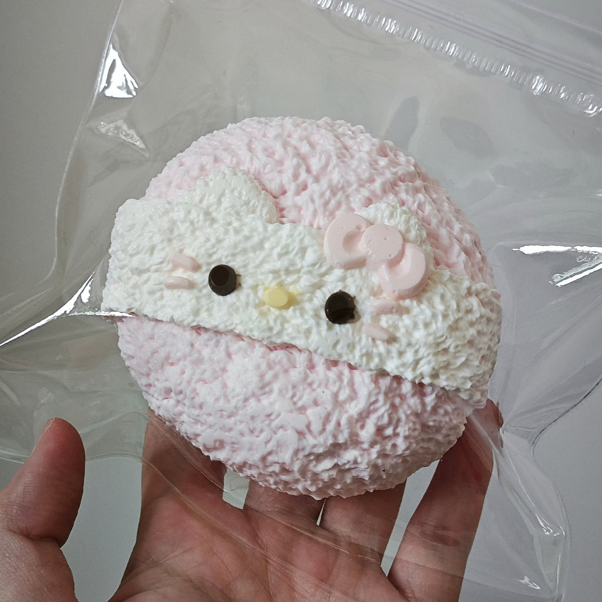 A hand holding a clear plastic bag containing a round, fuzzy pink and white Hello Kitty-themed item with embroidered facial details.