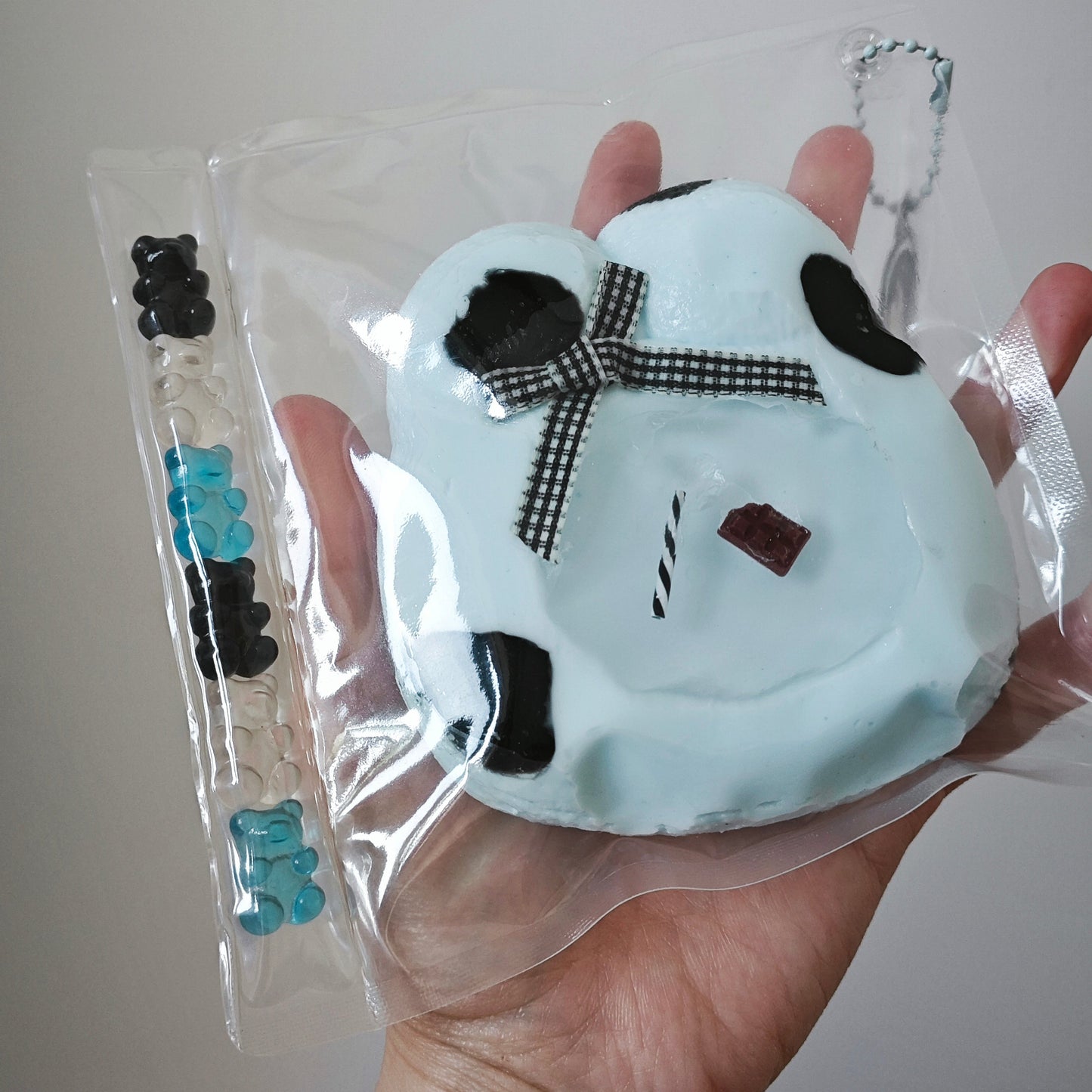 A hand holding a packaged squishy toy shaped like a panda face, with a pale blue body, black and white patches, a black-and-white bow, and small decorative details. A clear plastic bag with beads is also visible beside the main toy.