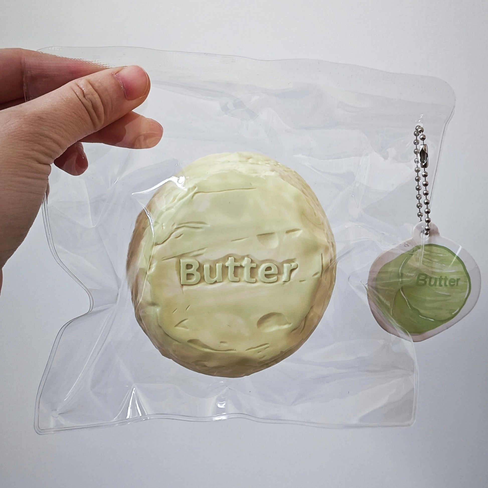 A hand holds a clear plastic bag containing a round, pale yellow-green novelty item labeled 'Butter,' with a smaller matching keychain or charm visible to the right on a plain light background.