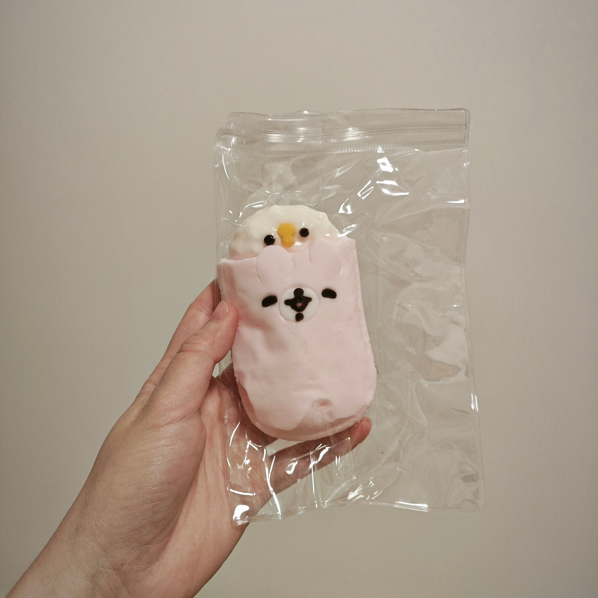 A hand holding a clear plastic bag containing a small plush toy shaped like a white bird peeking out from a pink pouch, against a plain beige wall.