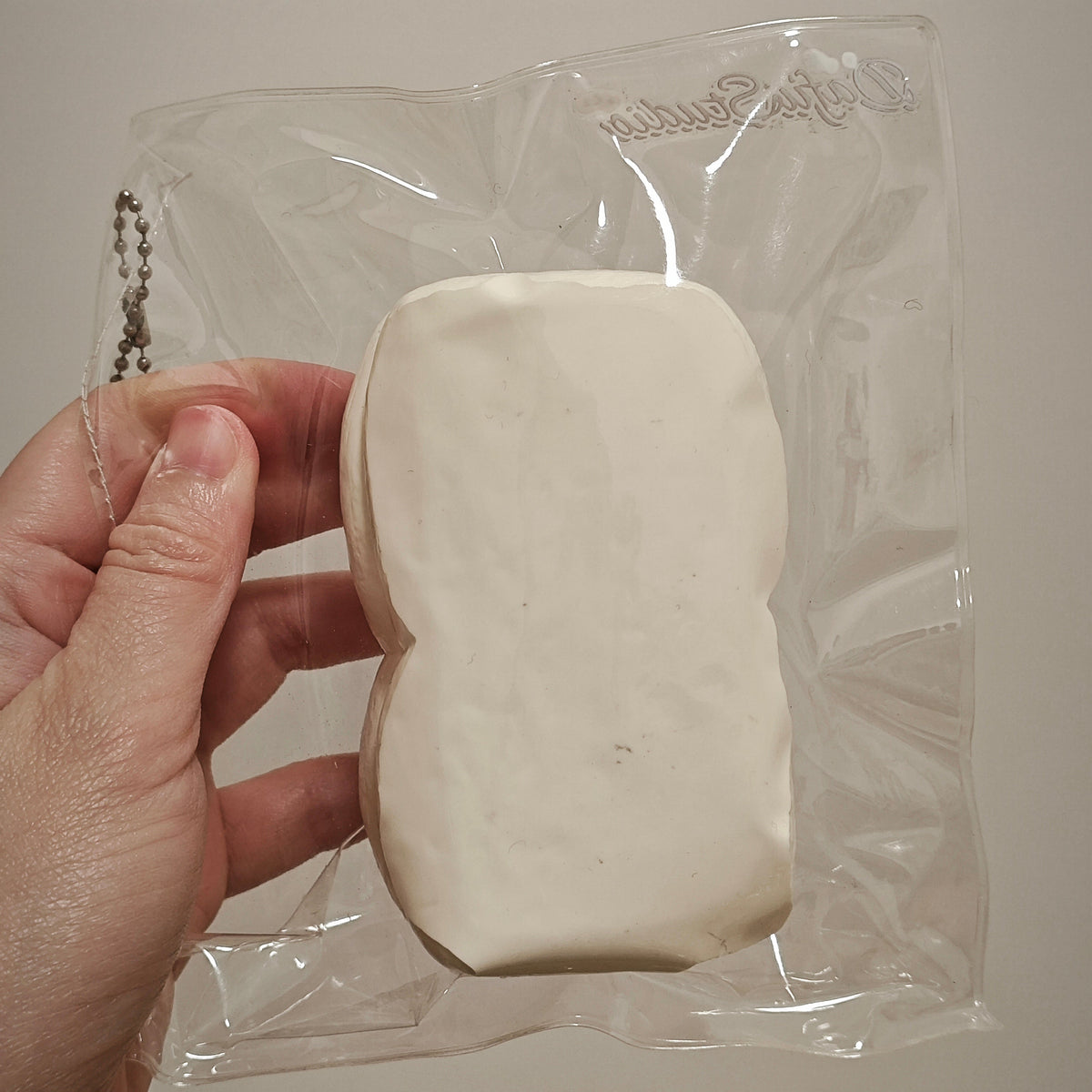 A hand holding a white, irregularly shaped foam or sponge-like item inside a clear plastic bag against a plain light beige background.