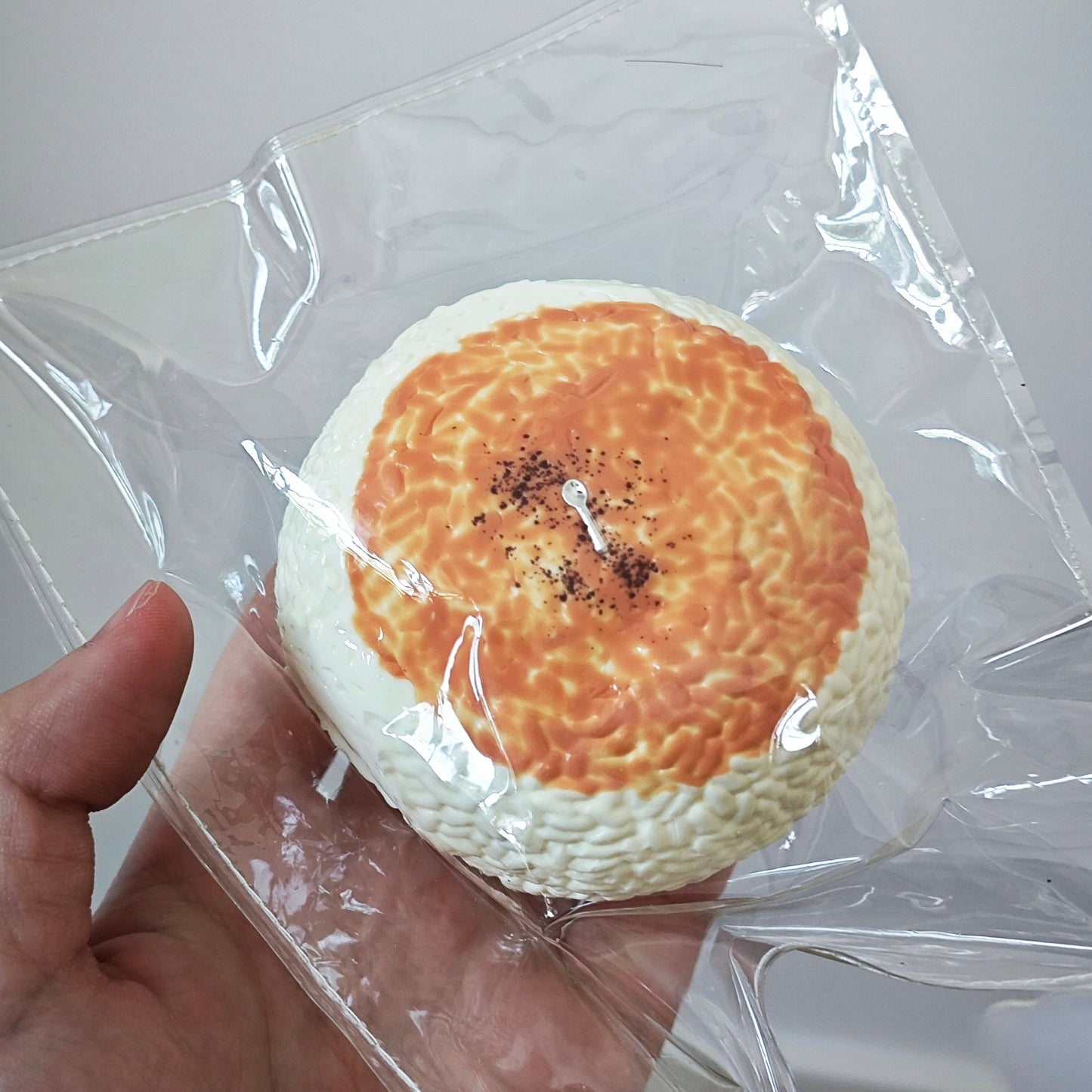 A hand holding a round, textured novelty item inside a clear plastic bag against a plain light gray background.