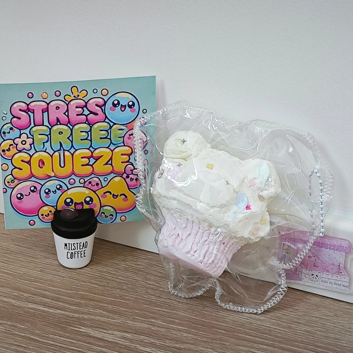A pastel pink cupcake-shaped purse wrapped in clear plastic sits on a light wood floor next to a colorful 'STRESS FREE SQUEEZE' card and a small black-and-white 'Mistral Coffee' cup. The background is a plain white wall.
