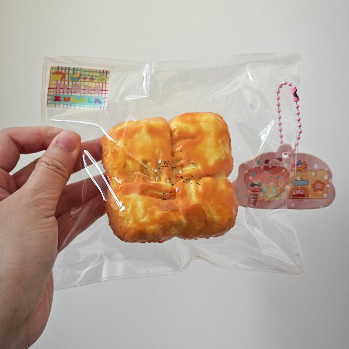 A hand holding a clear plastic bag containing a yellow-orange baked bread or pastry toy with small black seed-like details, alongside a pink cloud-shaped keychain charm with a pink chain.