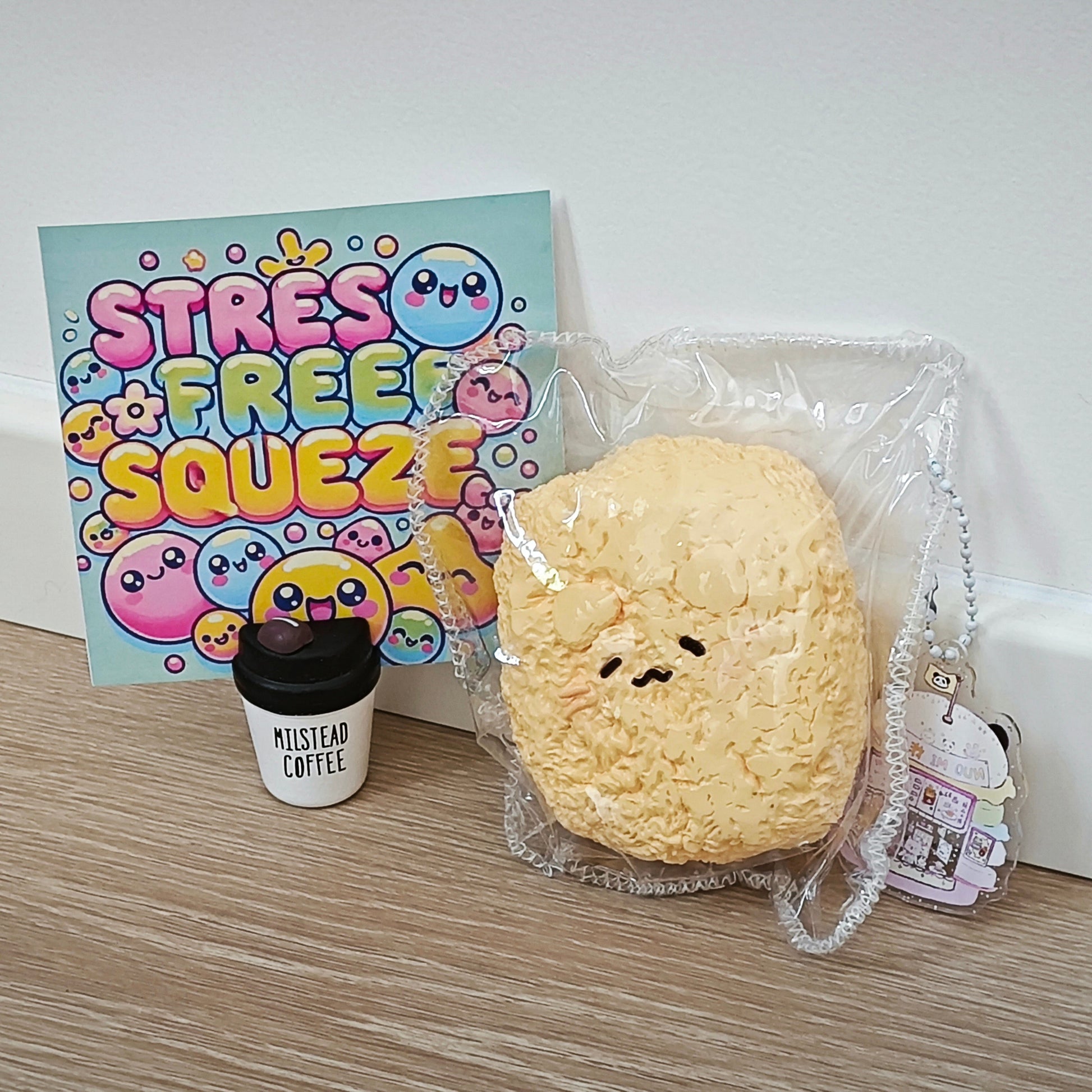 A cute yellow squishy toy shaped like a round character face, packaged in clear plastic, displayed next to a small black coffee cup and a colorful 'Stress Free Squeezie' card on a wooden surface.
