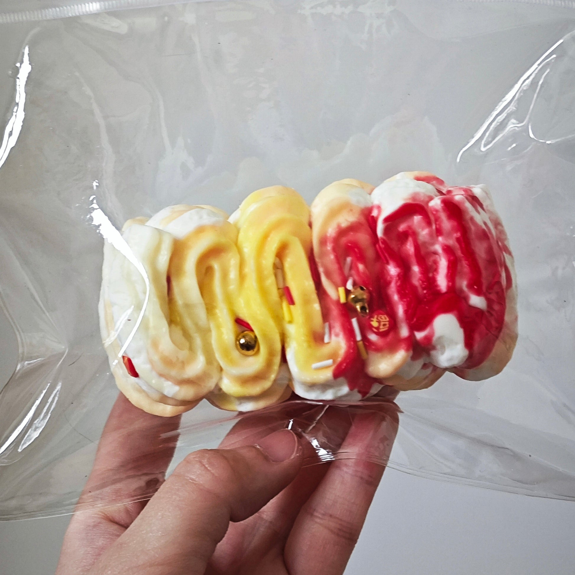 A hand holding a clear plastic bag containing a set of four novelty brain-shaped items, likely squishies or small decorative toys, in white, yellow, and red colors with small gold beads.