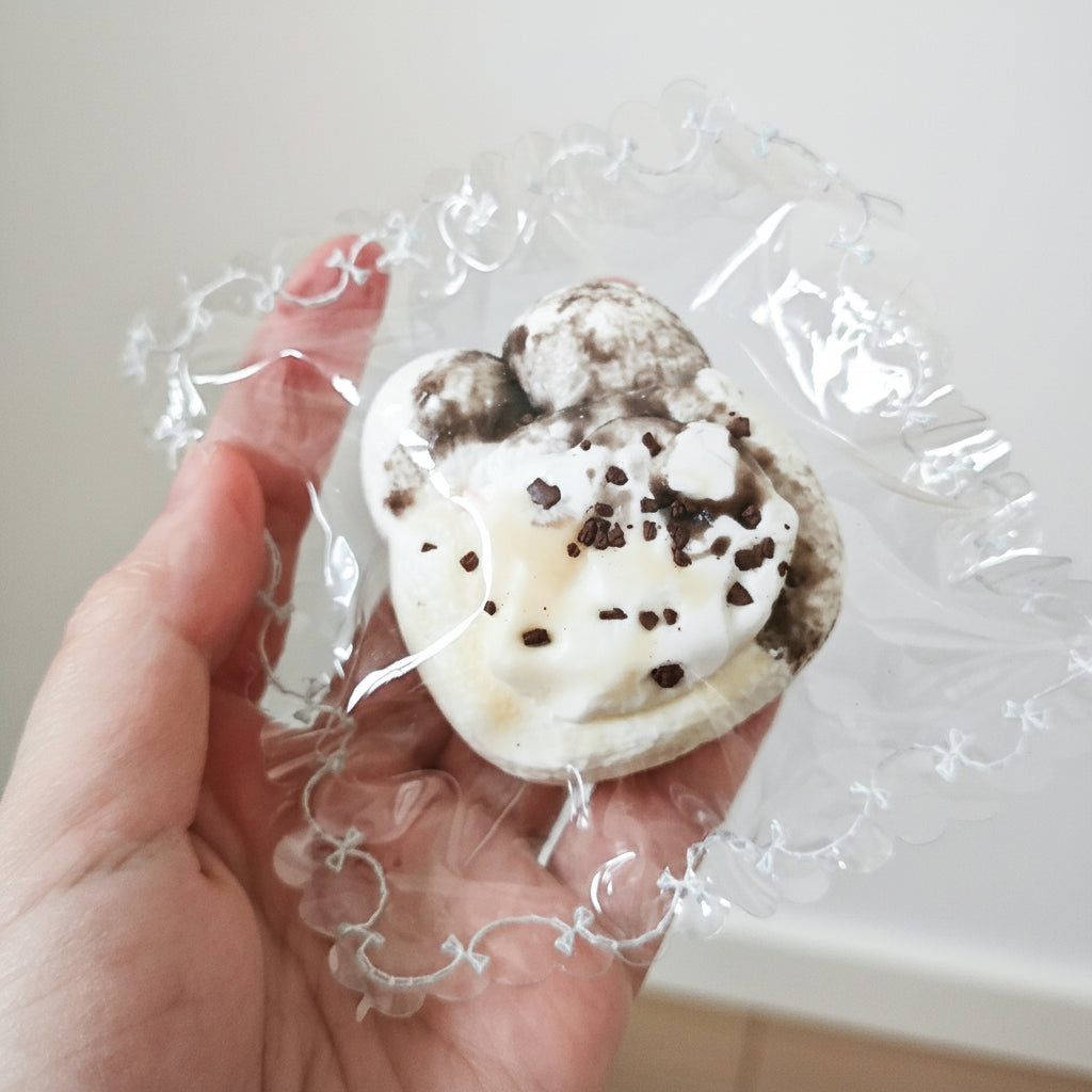 A hand holding a small, irregularly shaped white and dark brown cookie or candy piece wrapped in clear cellophane packaging against a plain indoor wall.