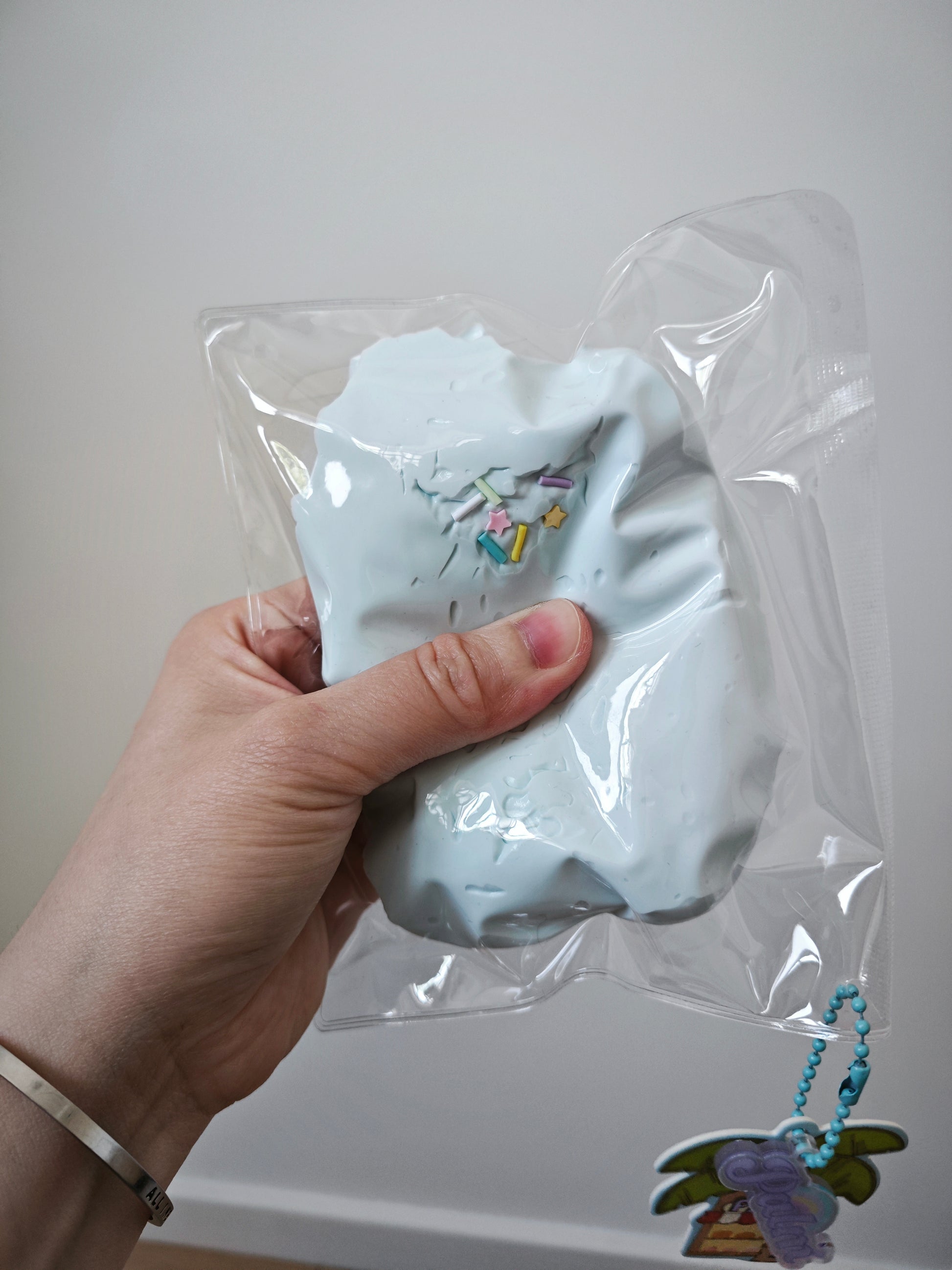 A hand holding a light blue squishy toy inside a clear plastic bag with a small hanging charm, against a plain light gray wall.