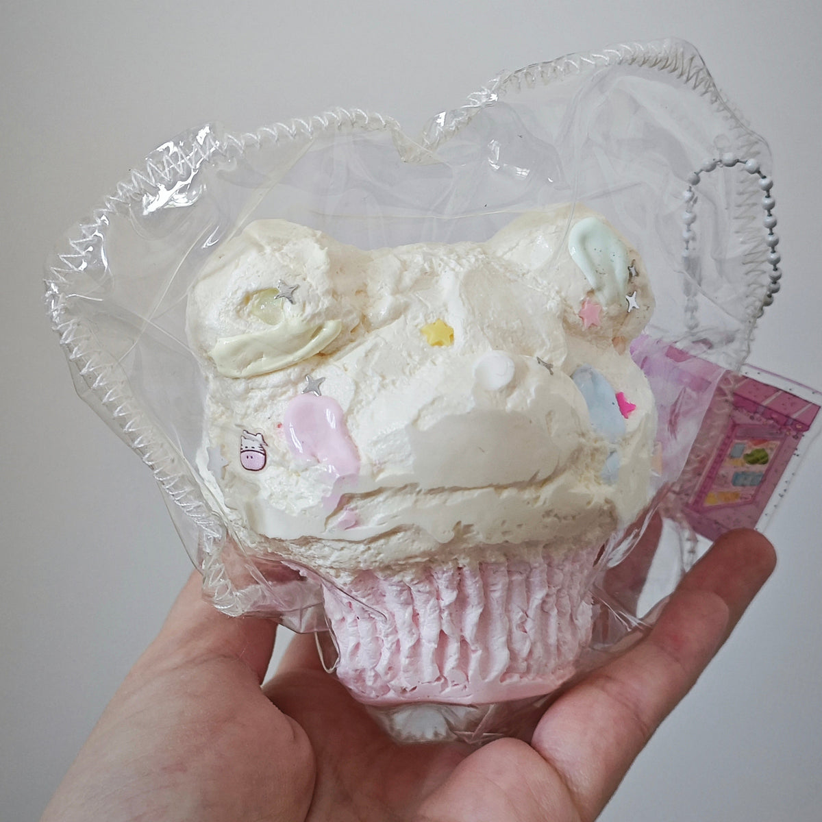 A hand holding a cupcake-shaped novelty item wrapped in clear plastic. The cupcake has off-white frosting with small pastel decorations and a pink wrapper at the bottom.
