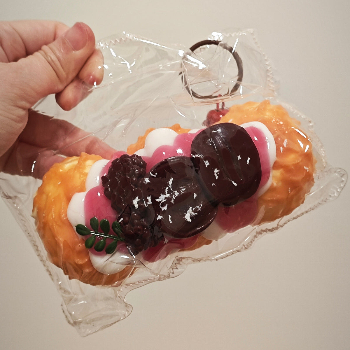 A hand holding a packaged novelty keychain shaped like a dessert, featuring orange pastry-like sides, pink and white cream details, dark brown chocolate drizzle, and a small green leaf accent, all inside a clear plastic bag.