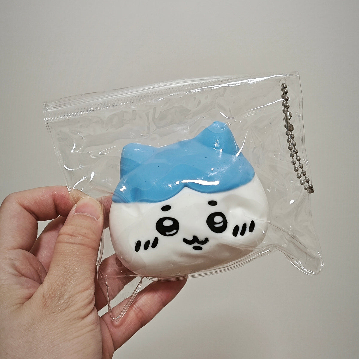 A hand holding a small, cute, cartoonish character squishy toy inside a clear plastic bag with a metal ball chain, against a plain light gray background.