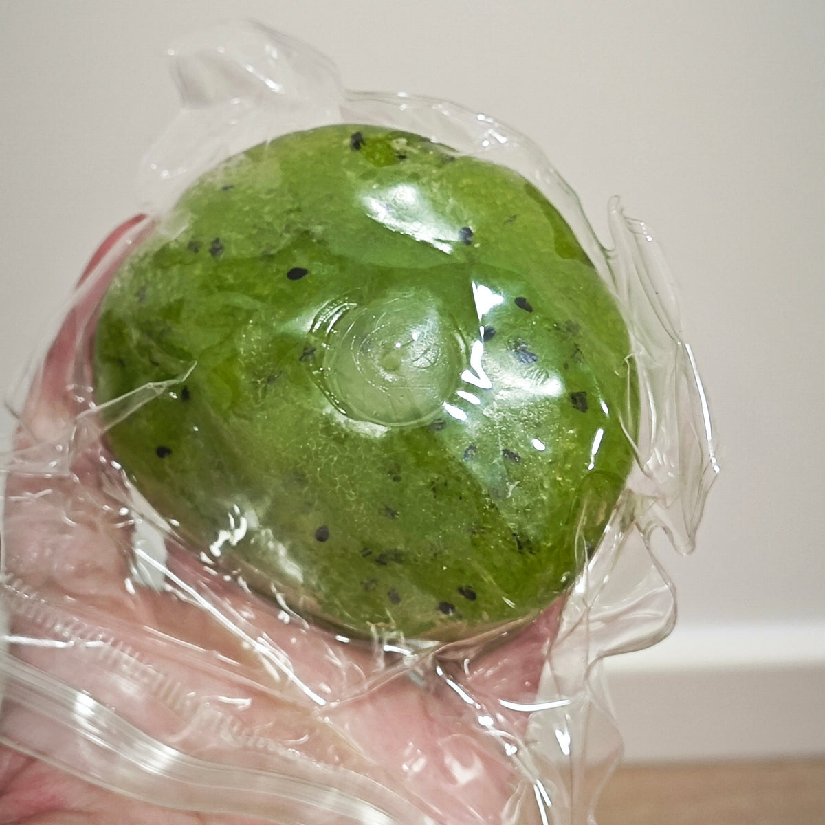 A green kiwi-shaped stress ball with black seeds, wrapped in clear plastic packaging and held in a hand.