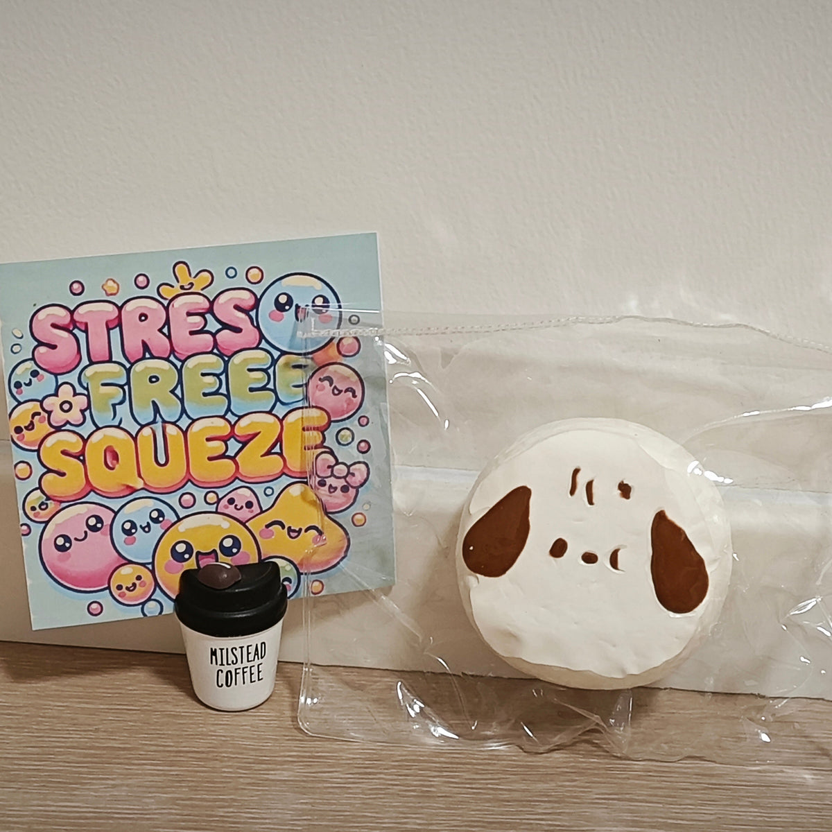 A cute dog-themed squishy toy in a clear plastic bag next to a colorful 'Stress Free Squeeze' card and a small coffee cup charm on a wooden surface against a plain wall.