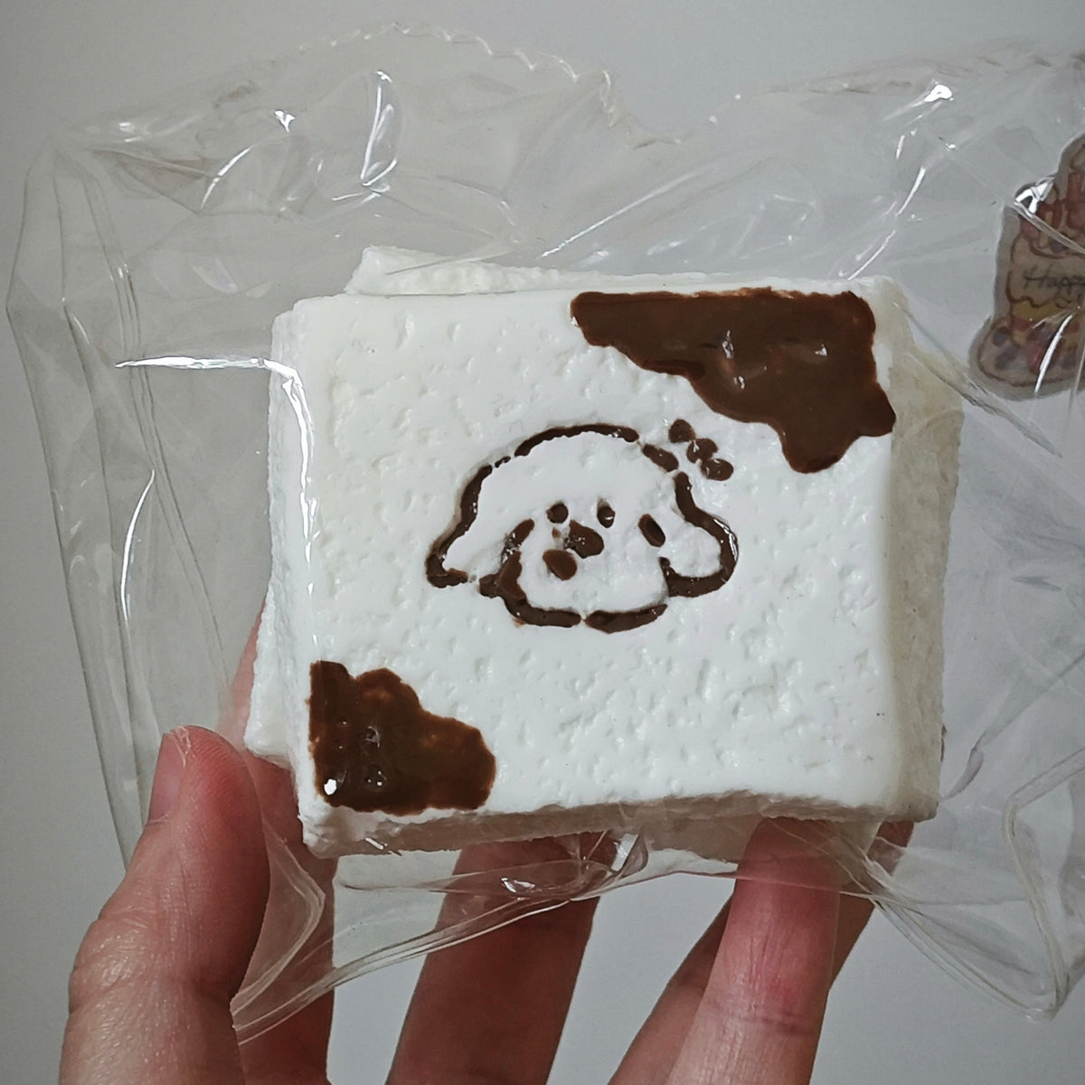 A hand holding a square white foam or sponge-like item wrapped in clear plastic, decorated with dark brown cow-print spots and a simple dog face illustration.