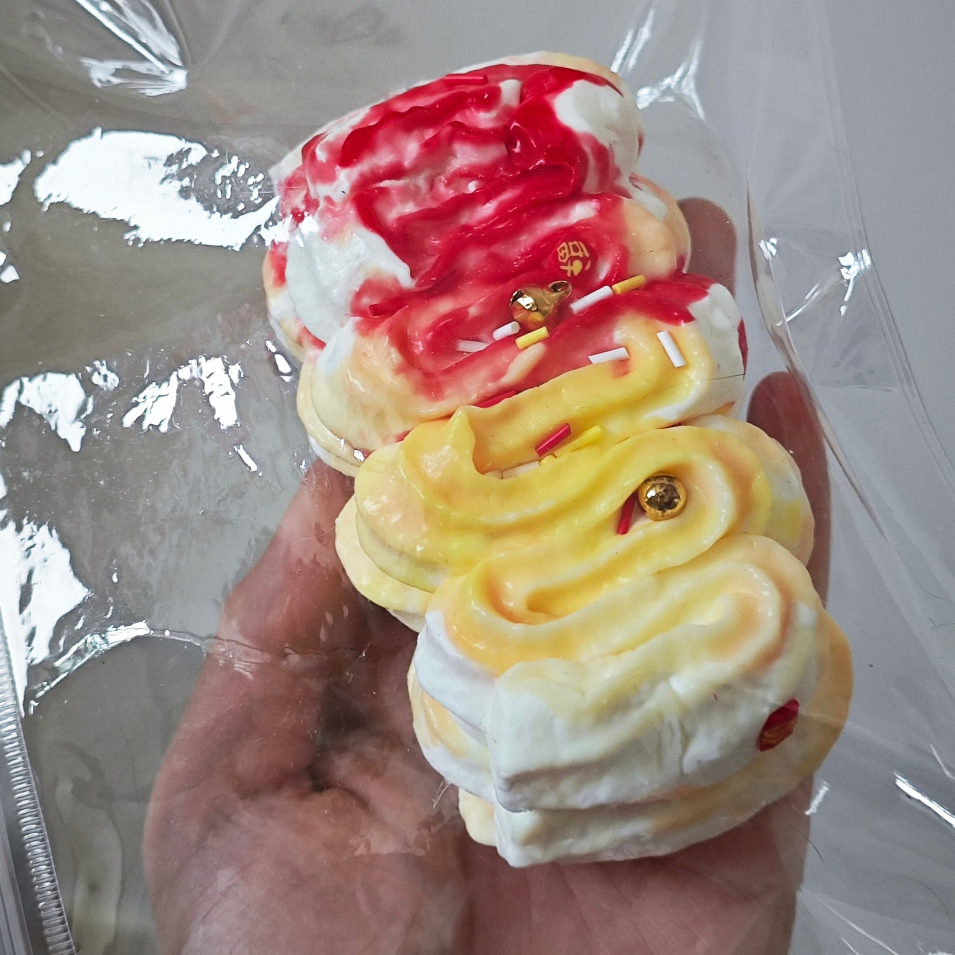 A hand holding a clear plastic bag containing three small, decorative, 3D-molded pastry-like pieces with swirls of red, yellow, and white frosting, and tiny gold bead accents.