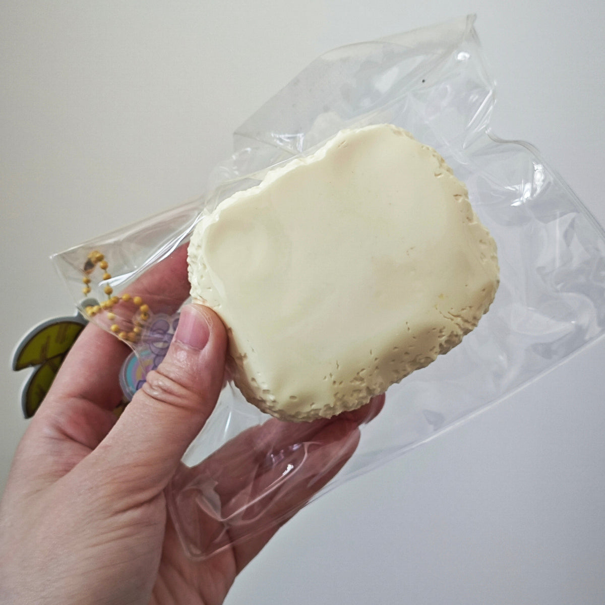 A hand holding a small, irregularly shaped cream-colored clay or resin piece inside a clear plastic bag against a plain light background.