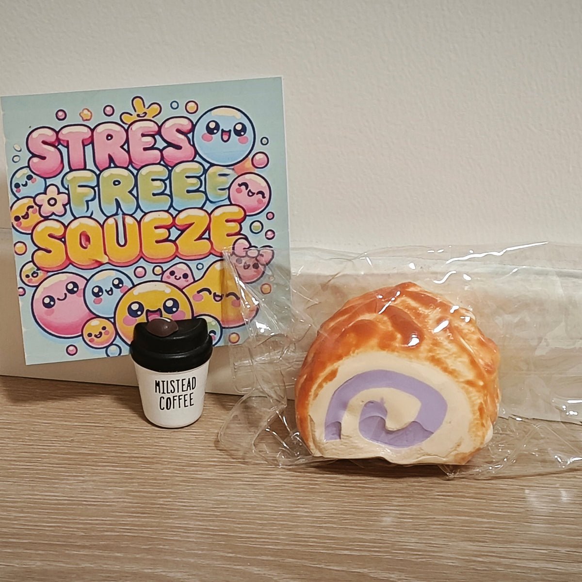 A packaged squishy toy shaped like a pastry roll with a purple swirl, displayed next to a small cup toy and a colorful 'Stress Free Squeeze' card on a wooden surface against a plain wall.