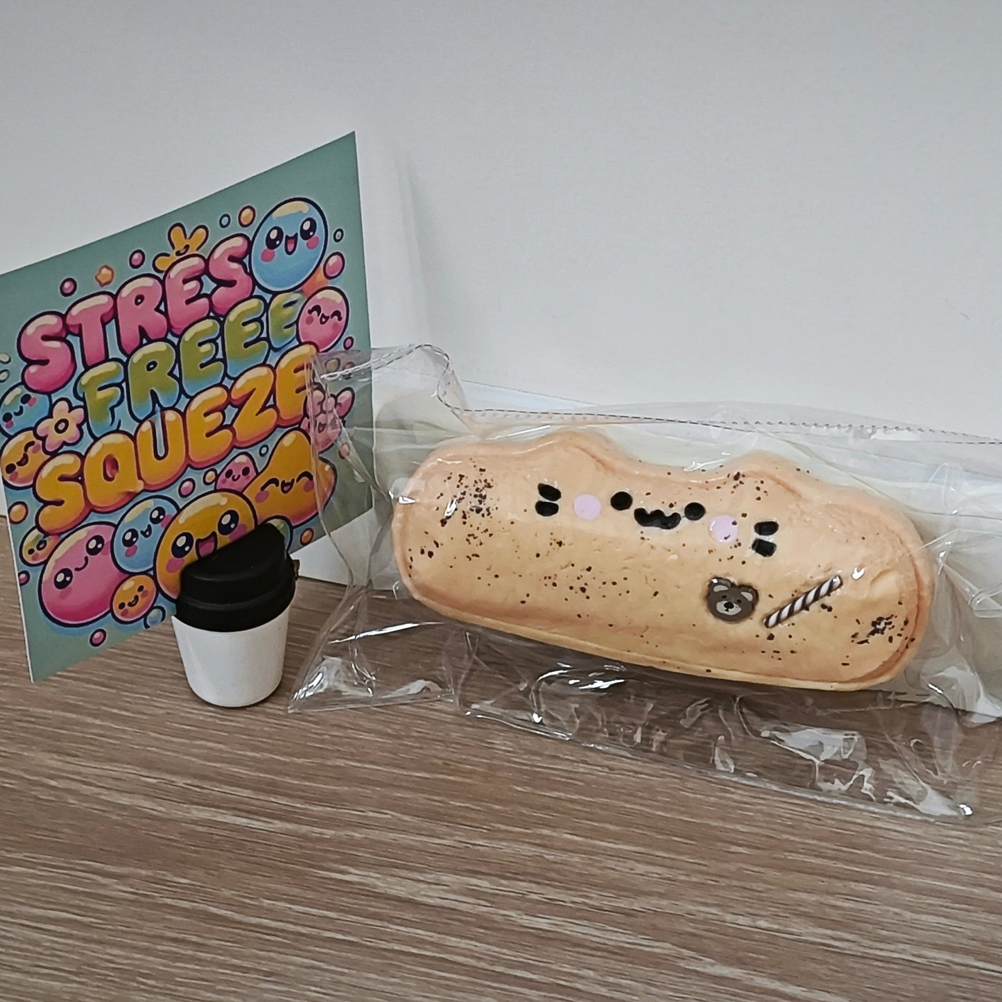 A packaged novelty stress toy shaped like a slice of toast or a pastry with a cute face, displayed on a wooden surface next to a small takeaway coffee cup and a colorful card that reads 'STRESS FREE SQUEEZE'.