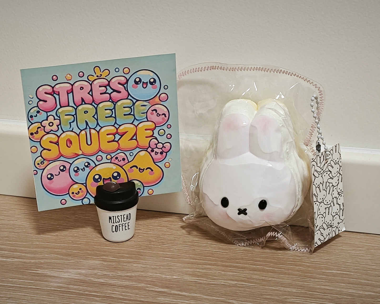 A white bunny-shaped squishy toy in clear plastic packaging sits on a wooden surface next to a colorful 'STRESS FREE SQUEEZE' card and a small black-and-white 'MILSTEAD COFFEE' cup. The background is a plain beige wall.
